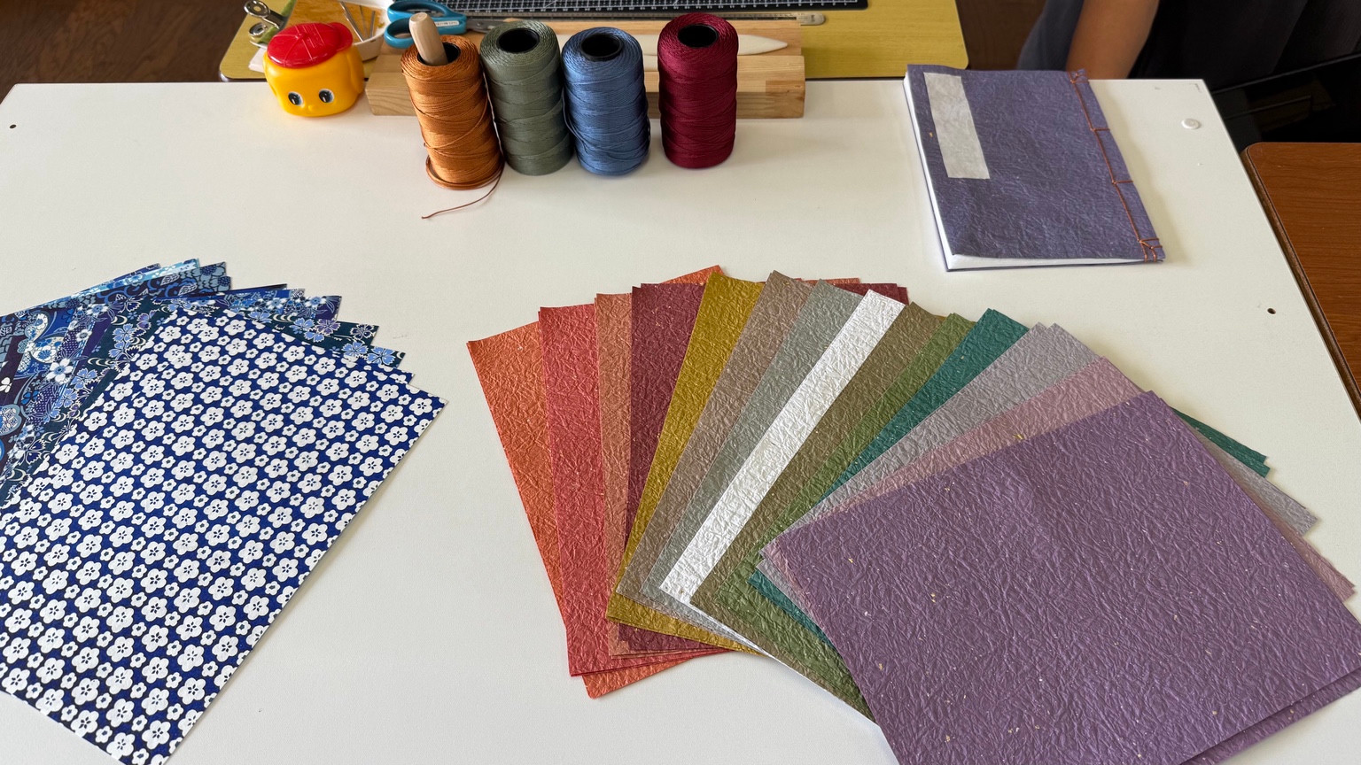 Colorful washi papers, binding threads and a sample handmade notebook arranged on a workshop table